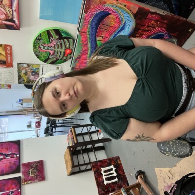 A photo of Melissa Brunner, a white curvy late 20's woman dressed in a dark green top and light denim shorts. Her head is slightly crooked, and she is wearing a pair of white headphones. Her background is of her art studio, decorated in paintings.