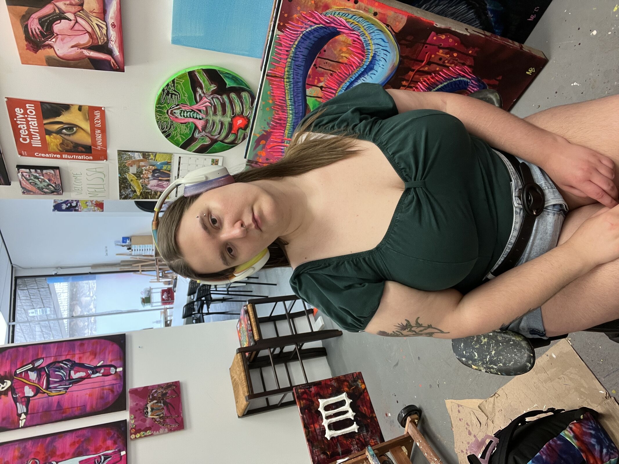 A photo of Melissa Brunner, a white curvy late 20's woman dressed in a dark green top and light denim shorts. Her head is slightly crooked, and she is wearing a pair of white headphones. Her background is of her art studio, decorated in paintings.